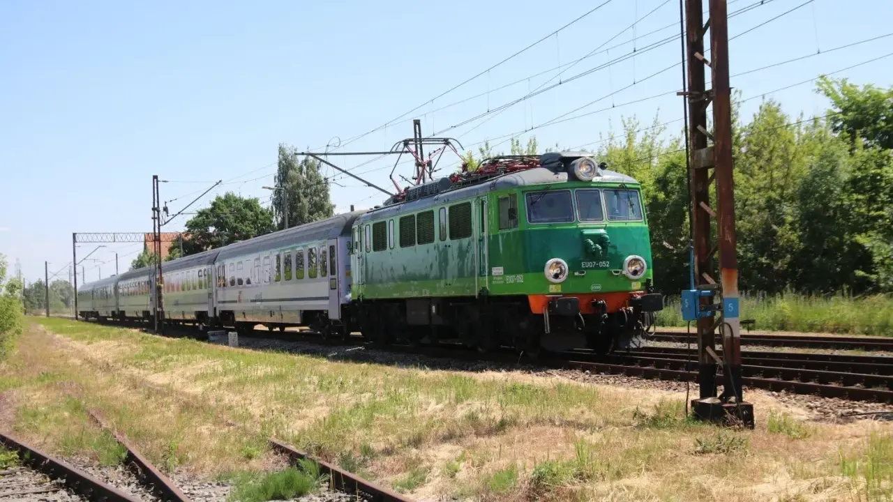 Zielony pociąg EU07-052 z wagonami mija tory w słoneczny dzień. To widok z wieluń trasa.