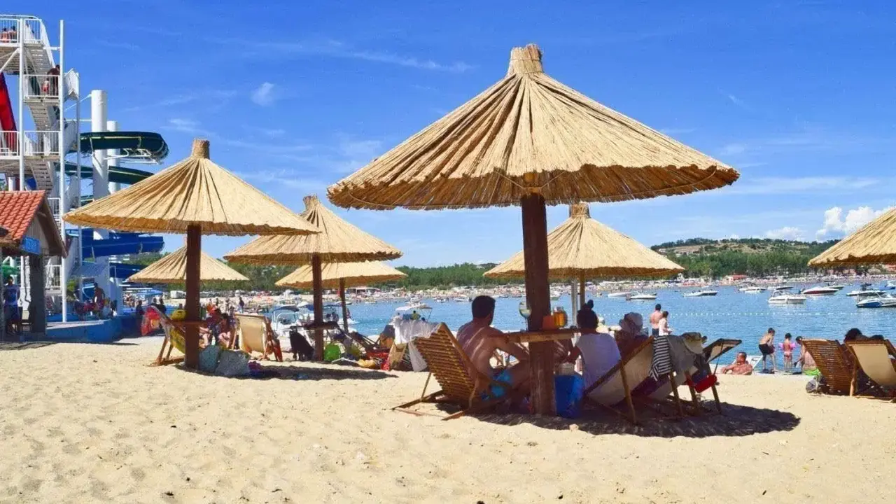 Słoneczna plaża na wyspie Rab, z ludźmi odpoczywającymi pod strzechami, w tle widać wodę z łodziami i zjeżdżalnie wodne.