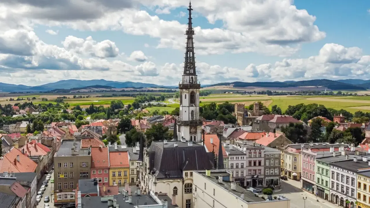Urokliwe miasto na trasie Wrocław, z dominującą wieżą ratuszową i kolorowymi kamienicami, otoczone zielonymi polami i wzgórzami.