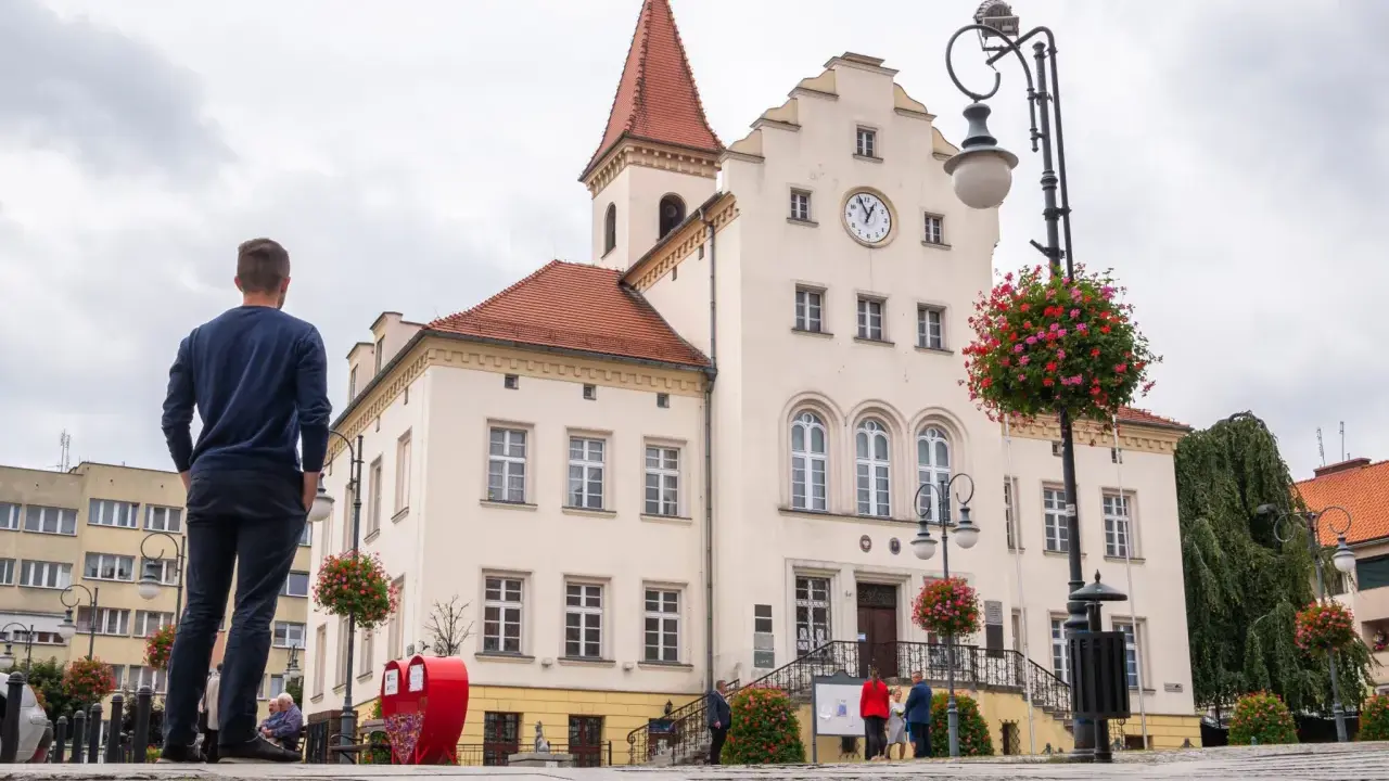 Mężczyzna stoi na rynku w Trzebnicy, podziwiając zabytkowy ratusz. Trasa spacerowa przez miasto prowadzi obok pięknych budynków.