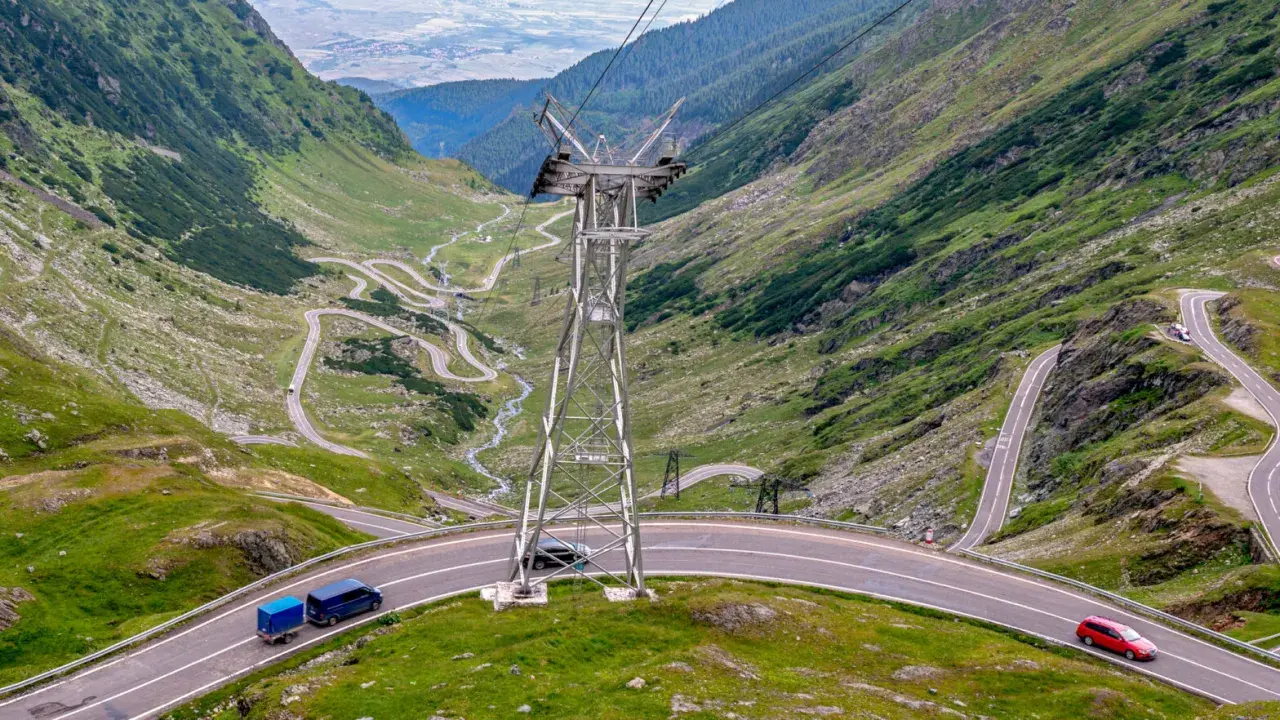 Kręte drogi w Rumunii wiją się przez zielone góry. Widać samochody i przyczepkę, a w oddali słup energetyczny.