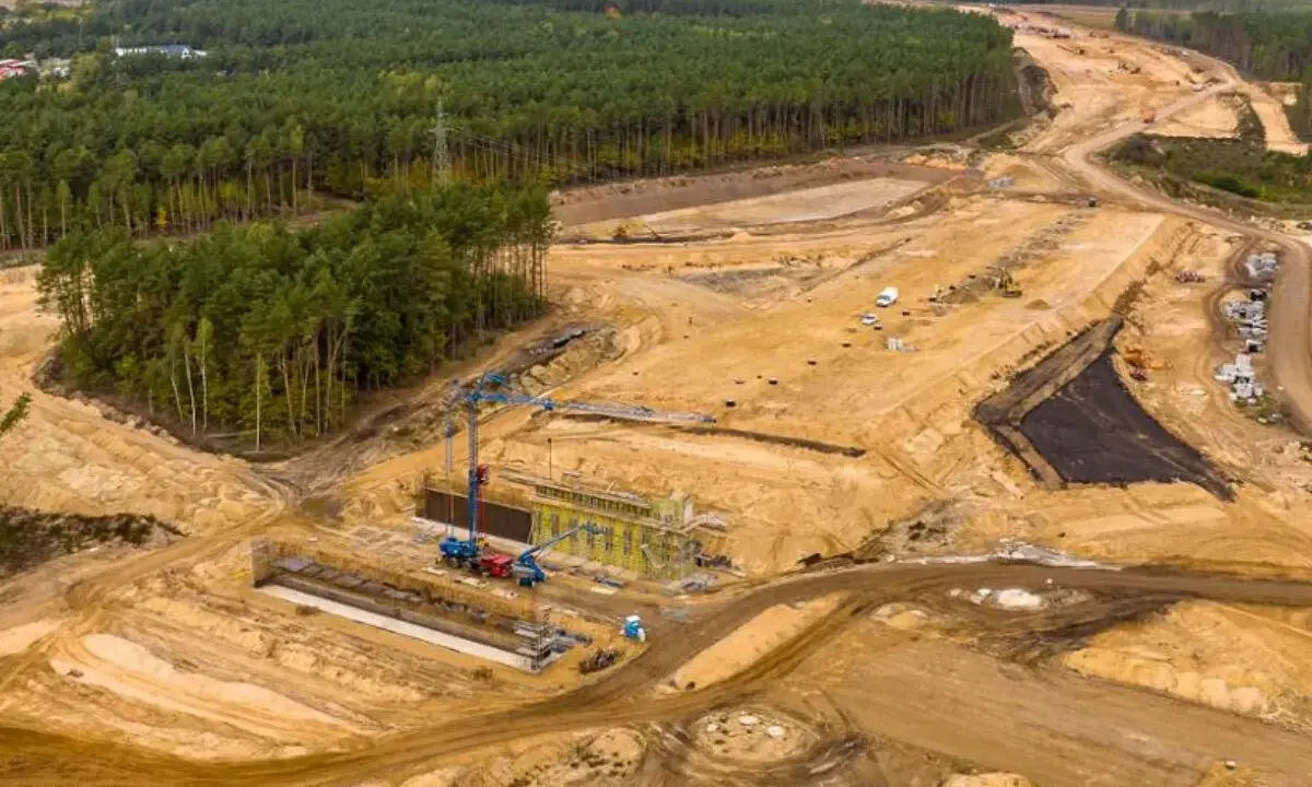 Ogromna budowa dróg w Polsce. Teren prac budowlanych z maszynami, dźwigami i przygotowywaną nawierzchnią, otoczony lasem.