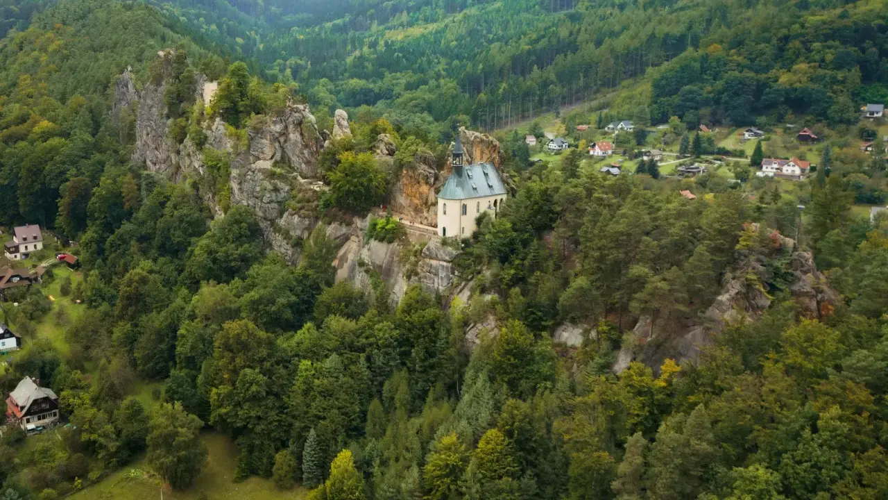 Malowniczy kościółek na skale, otoczony lasami, to jedno z **ciekawych miejsc w Czechach**. W oddali widać domki w dolinie.