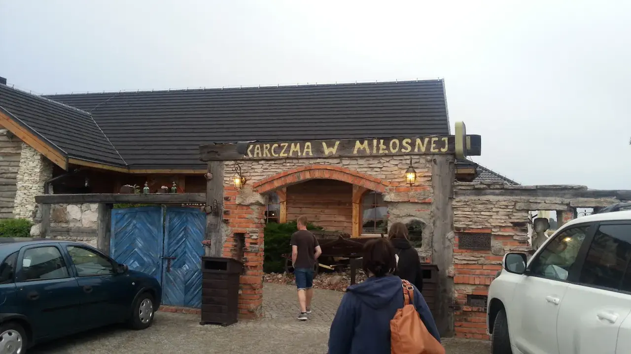 Karczma w Miłośnej, jedna z restauracji na trasie, zaprasza gości. Widać samochody i ludzi wchodzących do środka.