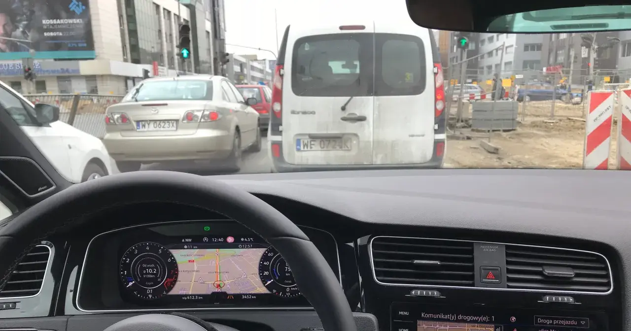 Utrudnienia na drodze, korek przed budową. Samochody stoją w deszczu, nawigacja pokazuje objazd.