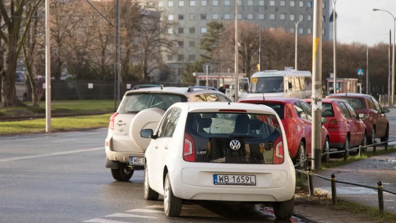 Biały Volkswagen up! zaparkowany tuż przy przejściu dla pieszych. W tle inne samochody i budynek.