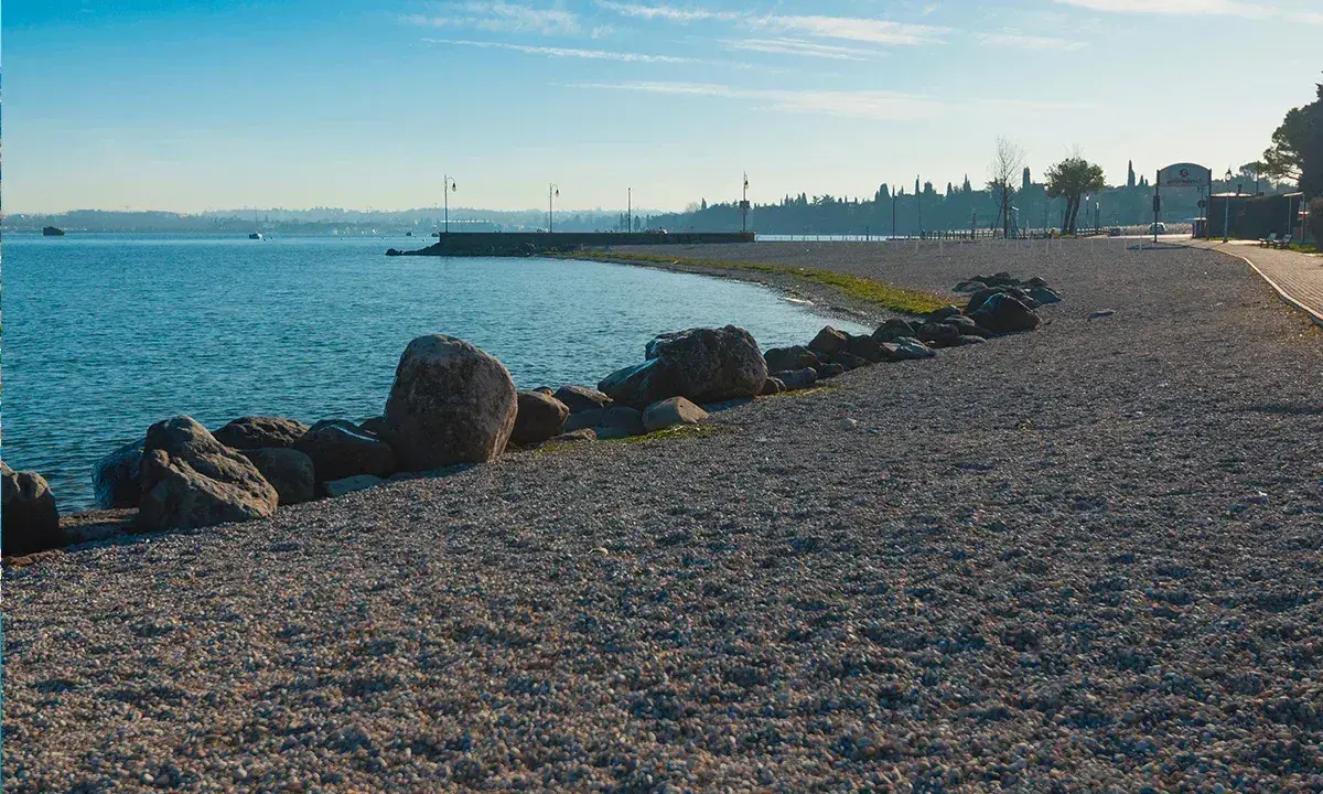 Kamienista plaża nad jeziorem, idealna na spacer po Peschiera del Garda. Widok na spokojną wodę i zarys miasta.