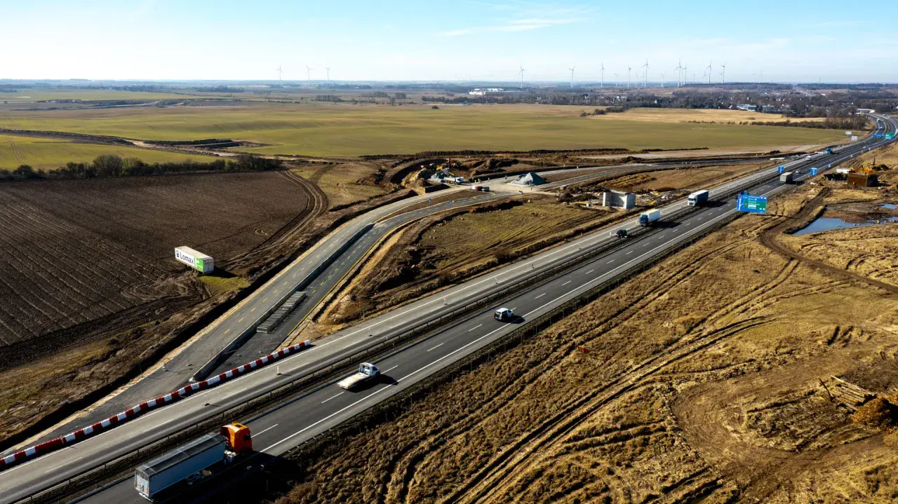 Rozbudowa autostrady A6 w otoczeniu pól i farm wiatrowych. Ruch samochodowy na ukończonych odcinkach, trwają prace budowlane.