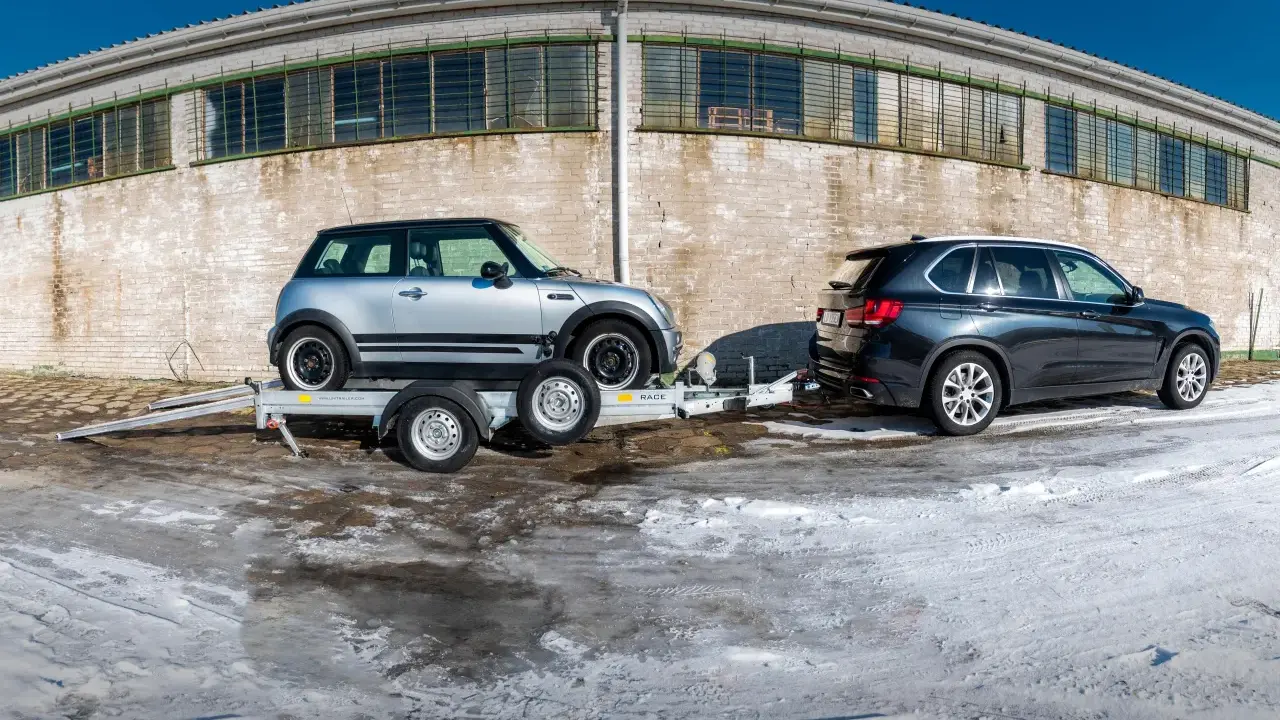 Srebrny Mini Cooper na lawecie, przygotowany do transportu. Obok stoi ciemny SUV. Mocowanie auta na lawecie jest kluczowe dla bezpiecznego przewozu.