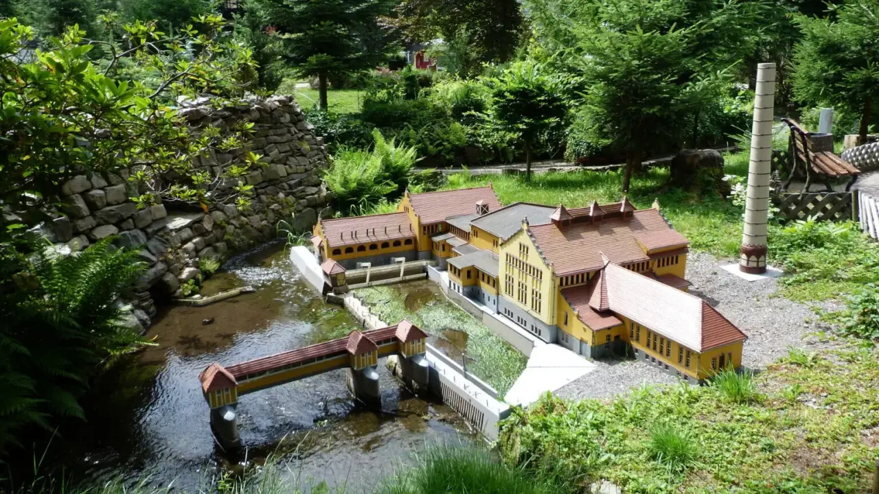 Miniaturowa zabudowa z mostkiem i rzeką, jak w parkach miniatur w Polsce. W tle drzewa i kamienny murek.