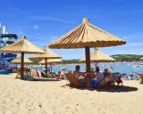 Słoneczna plaża na wyspie Rab, z ludźmi odpoczywającymi pod strzechami, w tle widać wodę z łodziami i zjeżdżalnie wodne.