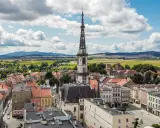 Urokliwe miasto na trasie Wrocław, z dominującą wieżą ratuszową i kolorowymi kamienicami, otoczone zielonymi polami i wzgórzami.