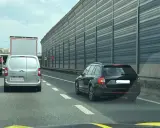 Czarny kombi jedzie pasem awaryjnym, ryzykując mandat. W tle inne pojazdy i ekrany akustyczne.