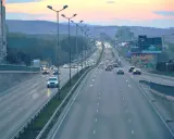Długa droga lokalna z wieloma samochodami w ruchu, oświetlona latarniami ulicznymi, z budynkami i wzgórzami w tle.