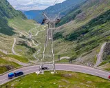 Kręte drogi w Rumunii wiją się przez zielone góry. Widać samochody i przyczepkę, a w oddali słup energetyczny.
