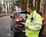 Policjant w odblaskowej kamizelce spisuje notatki przy zatrzymanym samochodzie. Może to być kontrola drogowa lub wystawianie mandatów w Czechach.