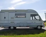 Biały ford galaxy camper z niebieskimi pasami stoi na poboczu drogi. W tle zielone pola i błękitne niebo.