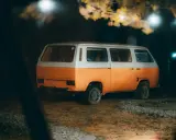 Pomarańczowo-biały kamper na nocnym parkingu, pod drzewami z lampkami. Czy można spać w kamperze na parkingu? Tak, to idealne miejsce na nocleg.