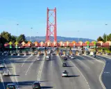 Ruchliwa autostrada w Portugalii z bramkami poboru opłat i charakterystycznym czerwonym mostem w tle.