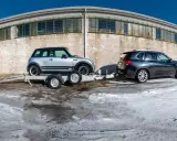 Srebrny Mini Cooper na lawecie, przygotowany do transportu. Obok stoi ciemny SUV. Mocowanie auta na lawecie jest kluczowe dla bezpiecznego przewozu.