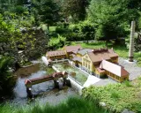 Miniaturowa zabudowa z mostkiem i rzeką, jak w parkach miniatur w Polsce. W tle drzewa i kamienny murek.