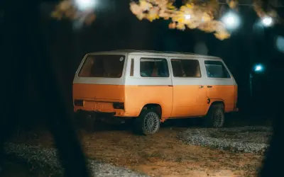Pomarańczowo-biały kamper na nocnym parkingu, pod drzewami z lampkami. Czy można spać w kamperze na parkingu? Tak, to idealne miejsce na nocleg.