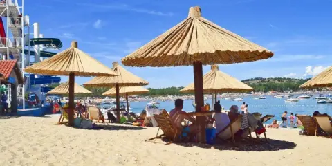 Słoneczna plaża na wyspie Rab, z ludźmi odpoczywającymi pod strzechami, w tle widać wodę z łodziami i zjeżdżalnie wodne.