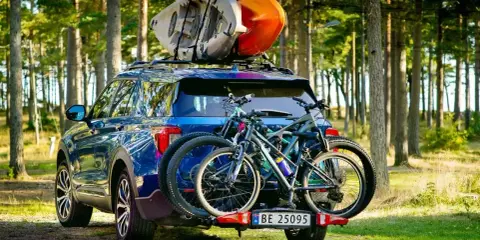 Niebieski SUV z dwoma rowerami na bagażniku i kajakami na dachu samochodu, gotowy na przygodę w lesie.