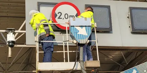 Pracownicy na podnośniku montują znak zakazu 2,0m, pokazując, jak działa e-toll.