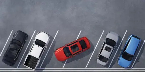 Czerwony samochód zajmuje dwa miejsca parkingowe, blokując sąsiednie pojazdy. Obok stoją czarny, biały pickup, srebrny i niebieski.