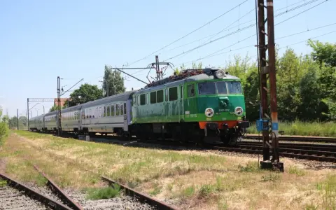 Zielony pociąg EU07-052 z wagonami mija tory w słoneczny dzień. To widok z wieluń trasa.