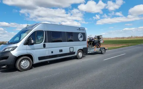 Srebrny kamper z motocyklem na przyczepie jedzie drogą. Przygoda w Szwecji kamperem dopiero się zaczyna.
