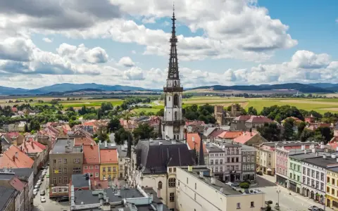 Urokliwe miasto na trasie Wrocław, z dominującą wieżą ratuszową i kolorowymi kamienicami, otoczone zielonymi polami i wzgórzami.