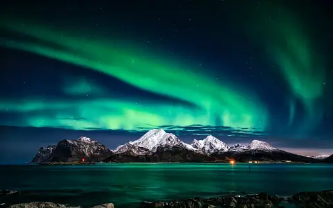 Zielona zorza polarna nad ośnieżonymi górami i morzem. Marzysz, jak dojechać do Norwegii, by zobaczyć to na żywo?
