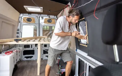 Mężczyzna buduje kamper z osobówki, montując okno. Widać drewniane elementy łóżka i instalację elektryczną.