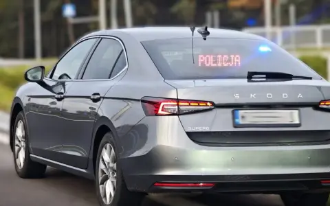 Szary, nieoznakowany radiowóz Skoda Superb z włączonymi niebieskimi światłami i napisem "POLICJA" na tylnej szybie.