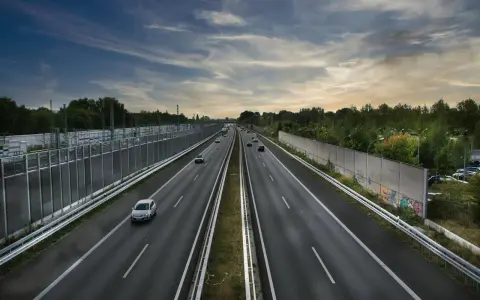 Szeroka autostrada z samochodami w oddali, otoczona zielenią i barierami dźwiękochłonnymi. Jazda autostradą pod pochmurnym niebem.