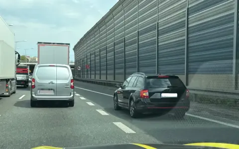 Czarny kombi jedzie pasem awaryjnym, ryzykując mandat. W tle inne pojazdy i ekrany akustyczne.