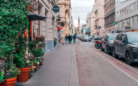 Wjazd do Wiednia samochodem osobowym. Ulica z kawiarniami, sklepami i zielenią, z zaparkowanymi autami i przechodniami.