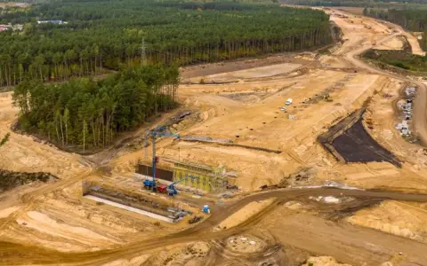 Ogromna budowa dróg w Polsce. Teren prac budowlanych z maszynami, dźwigami i przygotowywaną nawierzchnią, otoczony lasem.