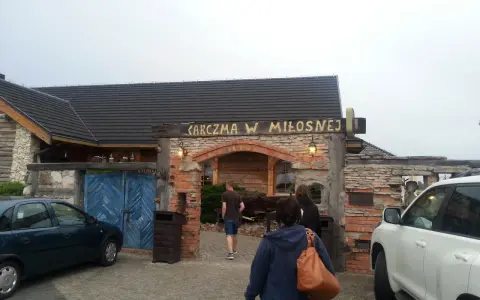 Karczma w Miłośnej, jedna z restauracji na trasie, zaprasza gości. Widać samochody i ludzi wchodzących do środka.