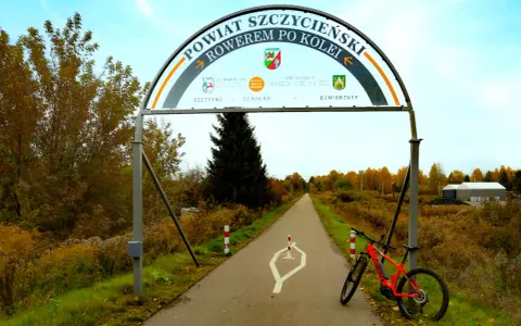 Pomarańczowy rower czeka na przygodę na trasie rowerowej w Szczytnie. Łuk nad drogą zaprasza do odkrywania powiatu.