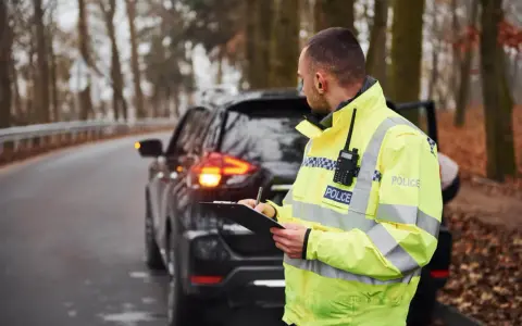 Policjant w odblaskowej kamizelce spisuje notatki przy zatrzymanym samochodzie. Może to być kontrola drogowa lub wystawianie mandatów w Czechach.