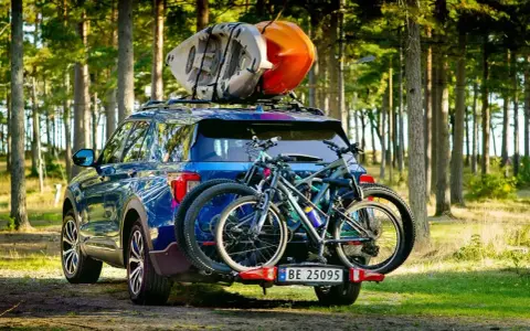 Niebieski SUV z dwoma rowerami na bagażniku i kajakami na dachu samochodu, gotowy na przygodę w lesie.