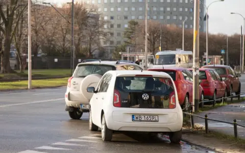 Biały Volkswagen up! zaparkowany tuż przy przejściu dla pieszych. W tle inne samochody i budynek.