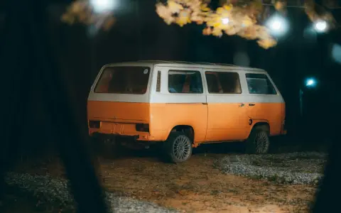 Pomarańczowo-biały kamper na nocnym parkingu, pod drzewami z lampkami. Czy można spać w kamperze na parkingu? Tak, to idealne miejsce na nocleg.