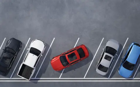 Czerwony samochód zajmuje dwa miejsca parkingowe, blokując sąsiednie pojazdy. Obok stoją czarny, biały pickup, srebrny i niebieski.