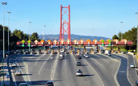 Ruchliwa autostrada w Portugalii z bramkami poboru opłat i charakterystycznym czerwonym mostem w tle.