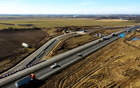 Rozbudowa autostrady A6 w otoczeniu pól i farm wiatrowych. Ruch samochodowy na ukończonych odcinkach, trwają prace budowlane.
