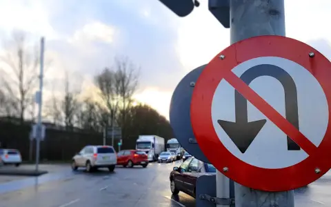 Znak zakazu zawracania informuje o nowych przepisach. Na mokrej drodze ruch samochodowy.